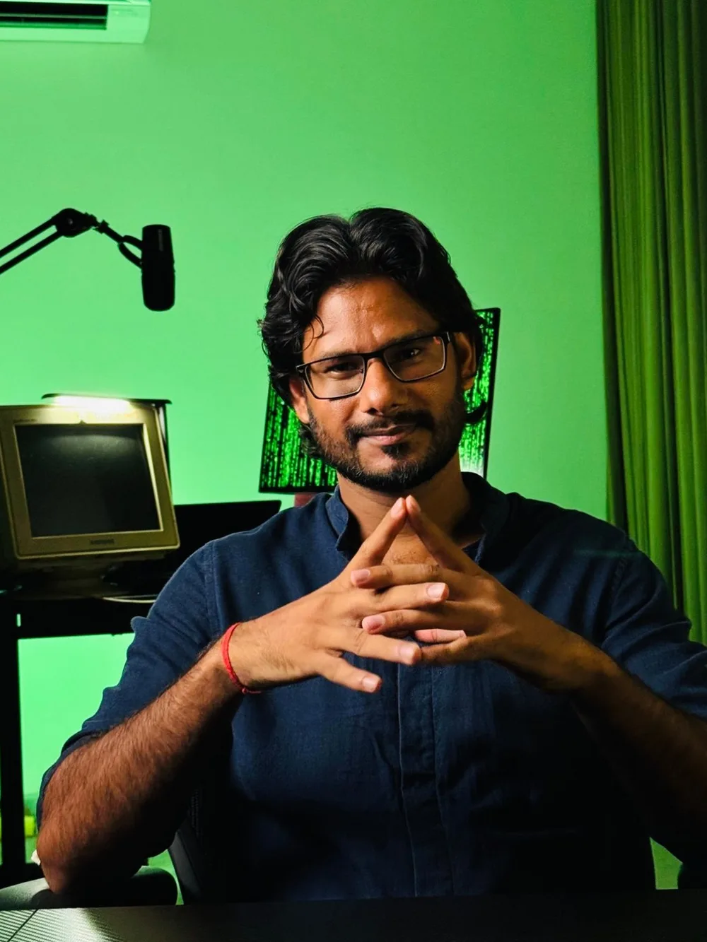 Gautam Kumawat at his studio desk, hands steepled, a boom microphone, a CRT monitor and a green-lit wall behind him